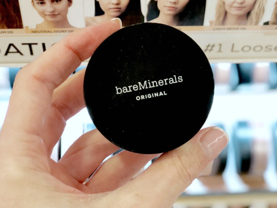 Hand holding a small black container of mineral based makeup in front of in-store display