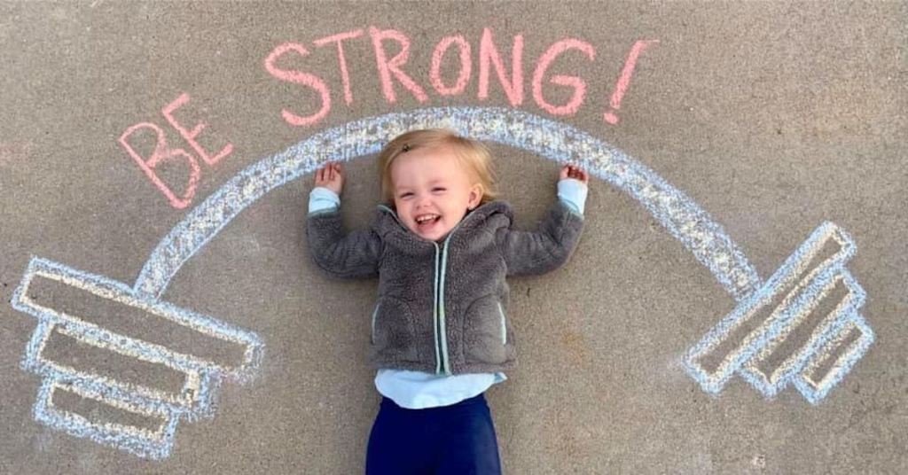 baby laying on sidewalk with chalk drawing of be strong and barbell weights
