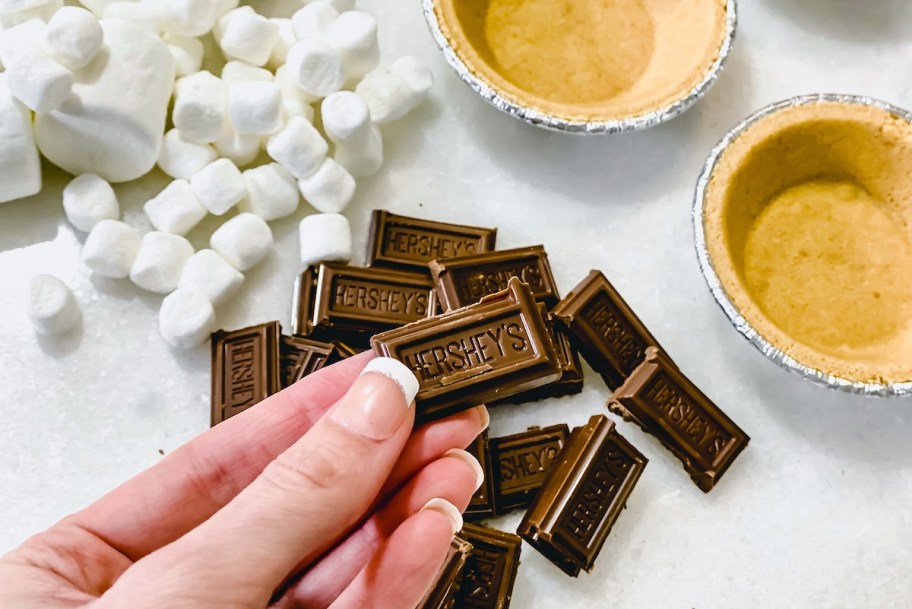 hand holding piece of hersheys chocolate with marshmallows and pie crusts on countertop