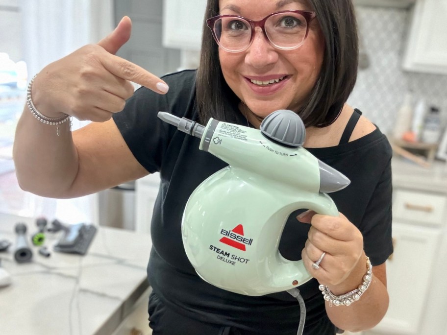 woman smiling and holding a green and grey Bissell hand steamer tool and pointing to