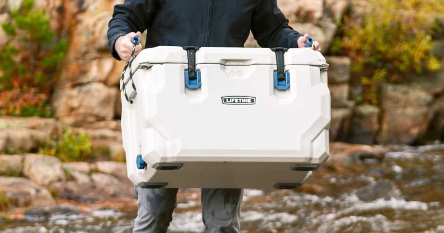 Person carrying a Lifetime 65-quart hard-sided cooler with blue latches and rope handles near a rocky stream.