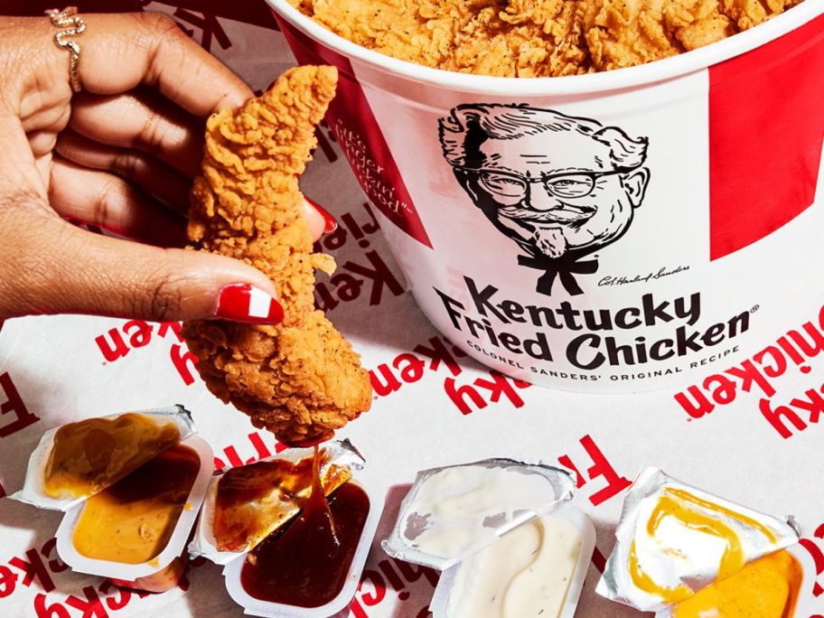 a hand with a KFC boneless tender about to dip it in a barbecue sauce container on a table with 3 other dipping sauces and a bucket of boneless tenders