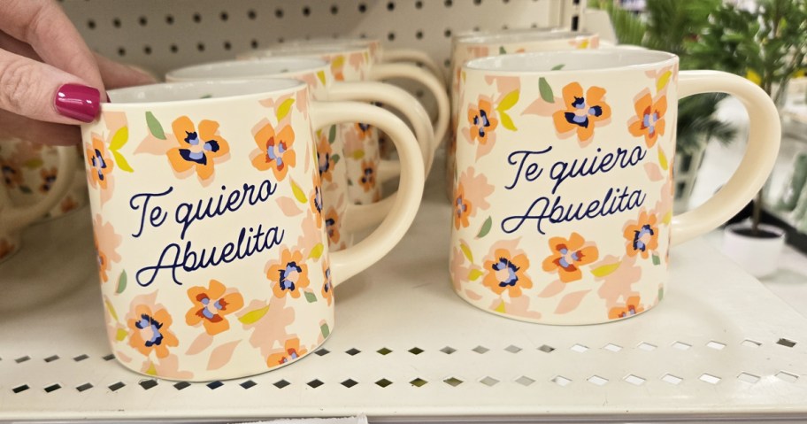 hand touching orange floral print mugs on shelf that say "Te Quiero Abuelita"
