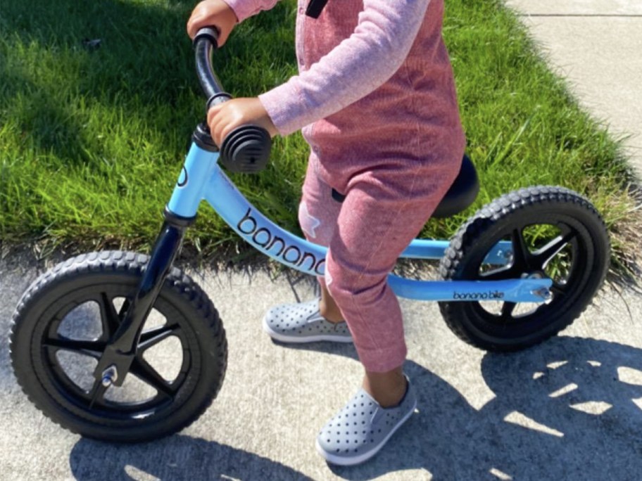 Little girl, riding blue banana balance bike, wearing a pink jumpsuit and gray shoes