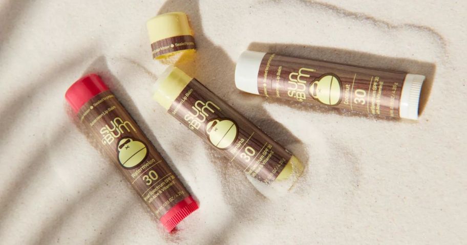 3 sun bum lip balms laying in sand, 2 open