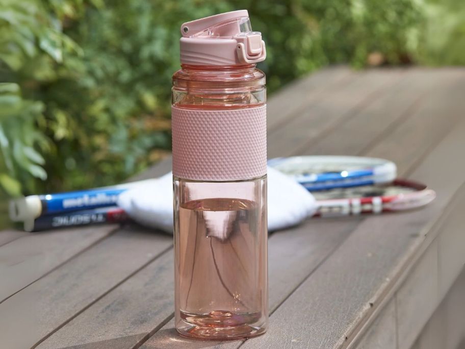 a pink water bottle on a wooden bench with tennis rackets in the background next it it