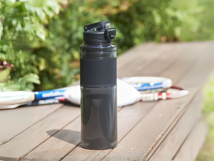 a black water bottle on a wooden bench with tennis rackets in the background next to it