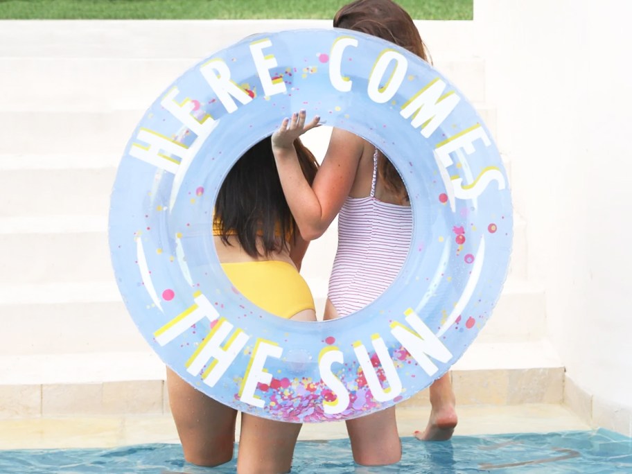 girls holding a pool tube that says "Here Comes The Sun"