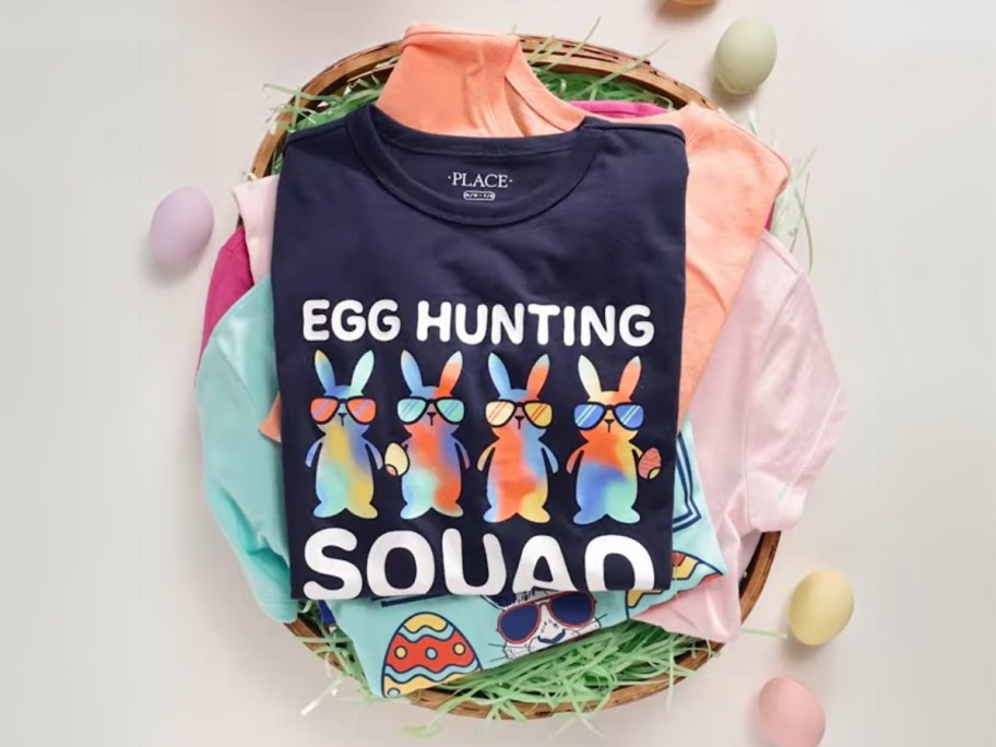 various kids Easter graphic tees stacked in an Easter basket with eggs around it