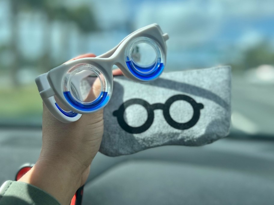 hand holding a pair of white and blue motion sickness glasses and a grey storage pouch 