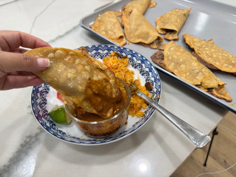 plated birria tacos being picked up and dipped into sauce next to a sheet pan with more tacos