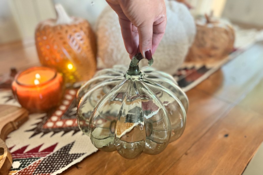 fingers picking up a green glass pumpkin by the stem with more fall decor in the background