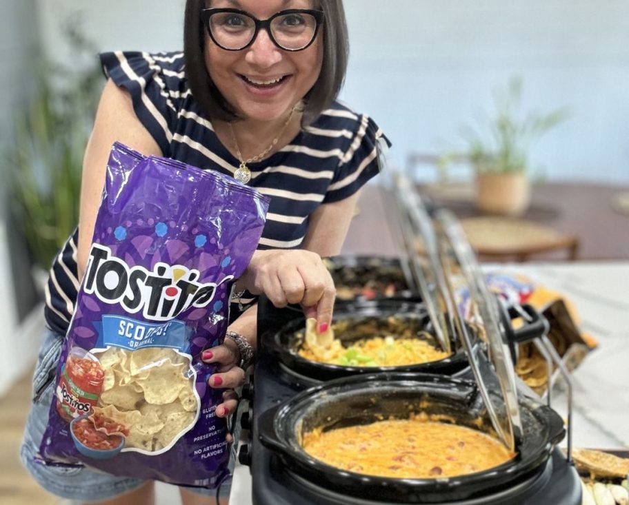 woman holding a bag of Tostitos and dipping a chip into one of the compartments of a Bella Triple Sow Cooker filled with 3 different dips