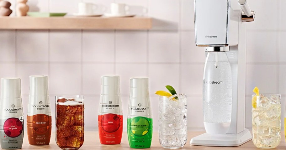 SodaStream flavors bottles and a glass of carbonated water with fruit slices next to a white SodaStream machine on a counter