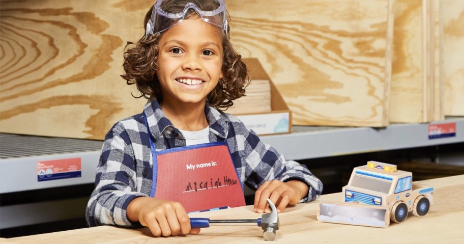 Child in workshop holding hammer next to toy snowplow.