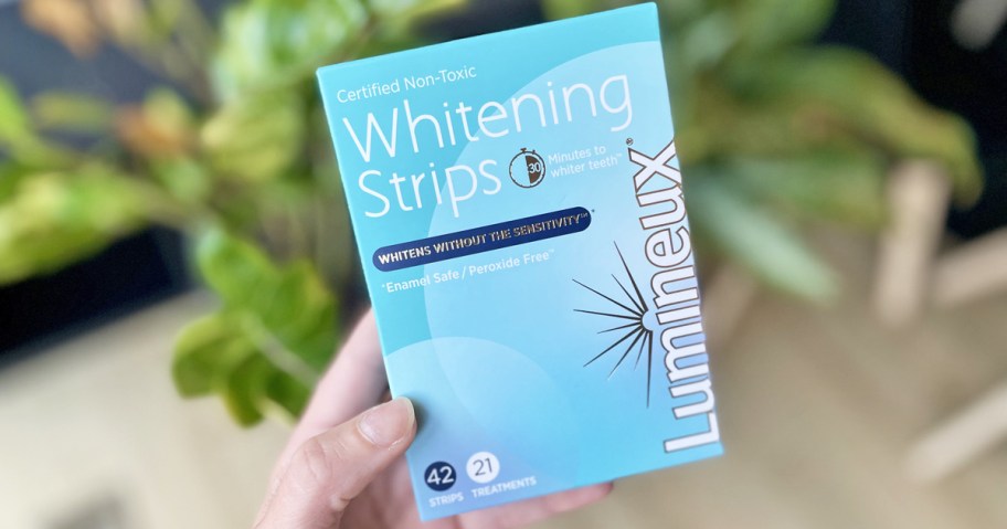 holding a large blue box of Lumineux Teeth Whitening Strips