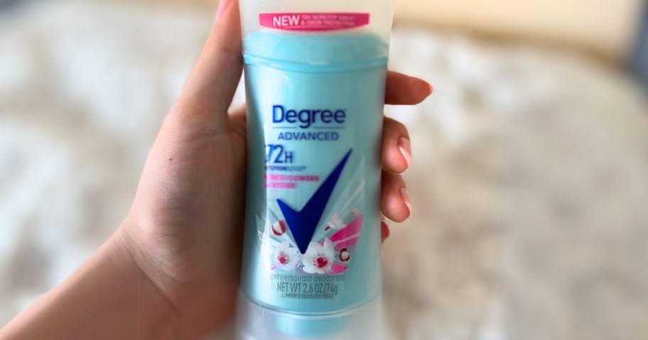hand holding a blue Degree Advanced Protection Antiperspirant Deodorant, white blanket can be seen over a bed behind it