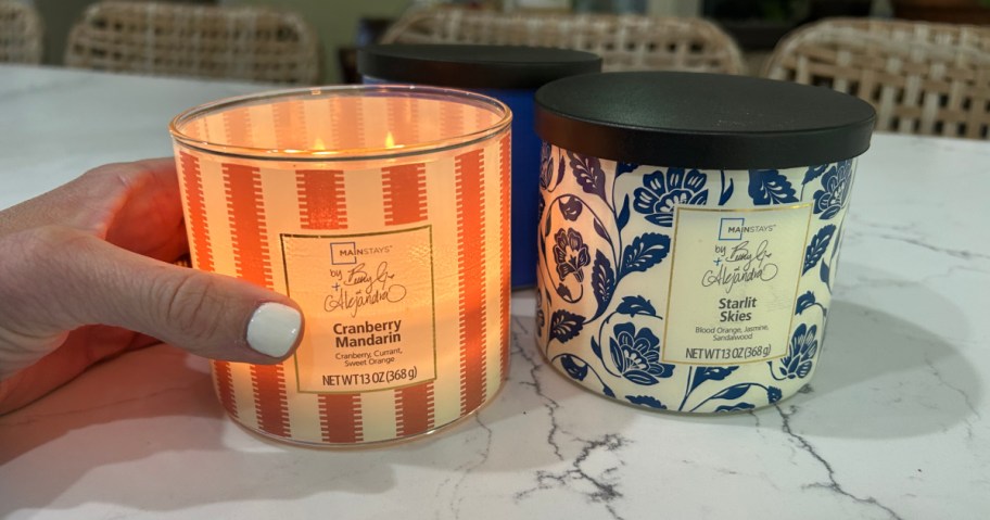 Person with white nail polish, holding orange and white striped candle next to blue floral patterned candle