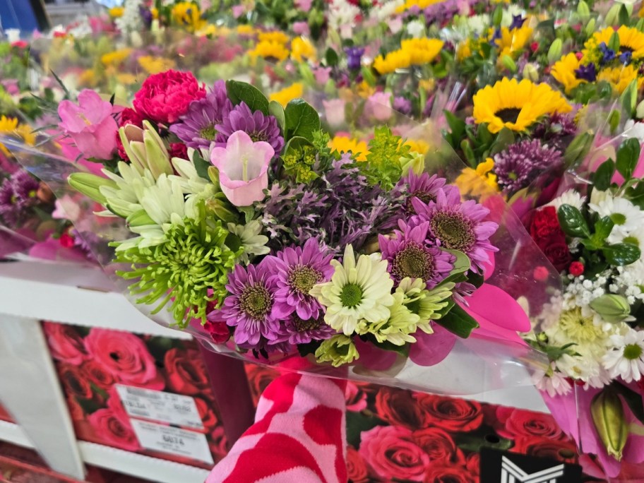 hand holding a bouquet of flowers with pink, purple and white tulips, daisies and other flowers wrapped in plastic - a display with more flower bouquets is behind it