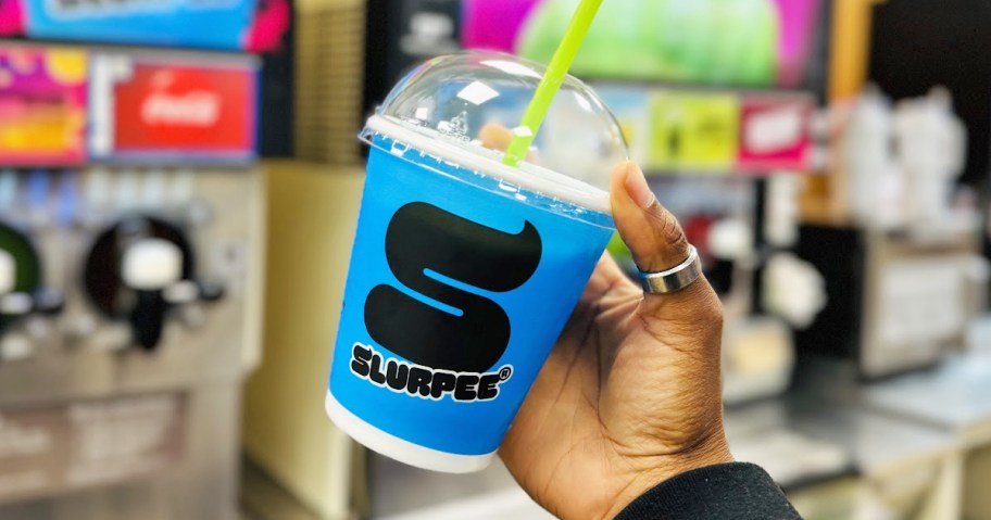 hand holding up a blue Slurpee cup in front of Slurpee machines