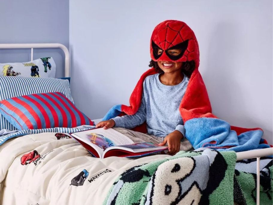 Child sitting on a bed made with Pillowfort Marvel bedding while wearing a Spiderman Hooded Throw Blanket