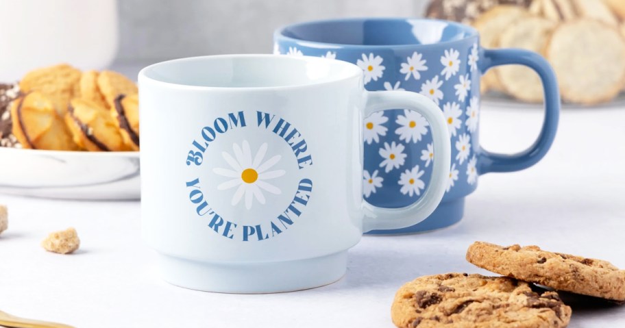 blue coffee mug that says "bloom where you're planted" and a darker blue mug with daisy print