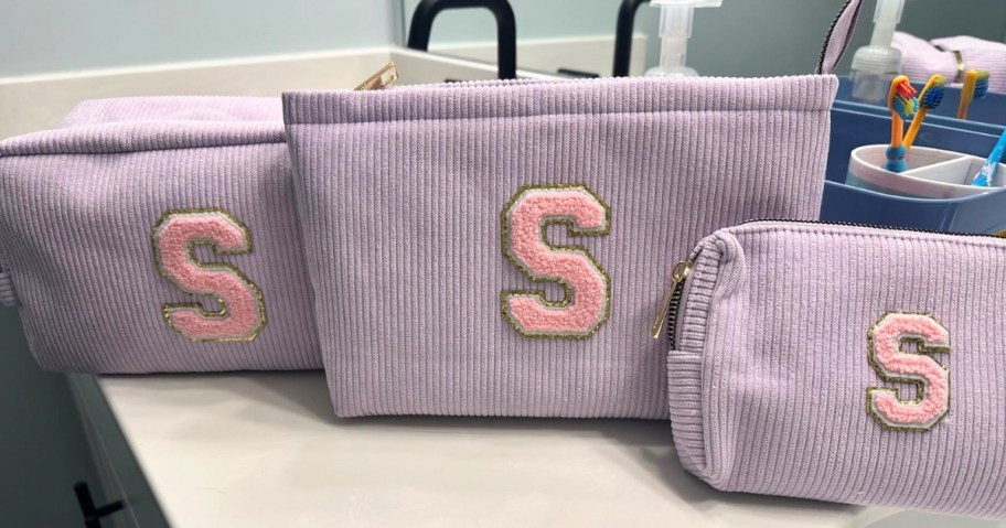 3 purple makeup bags with pink initials on them on a bathroom counter
