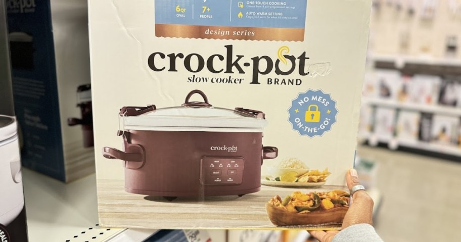 a reddish color CrockPot in a box on with a woman's hand pulling it off a store shelf