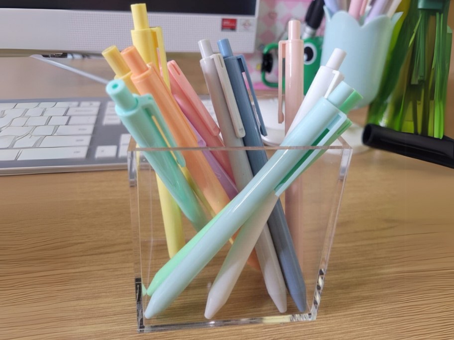 12 pastel color ink pens in a clear pencil cup, a computer and keyboard are behind it 