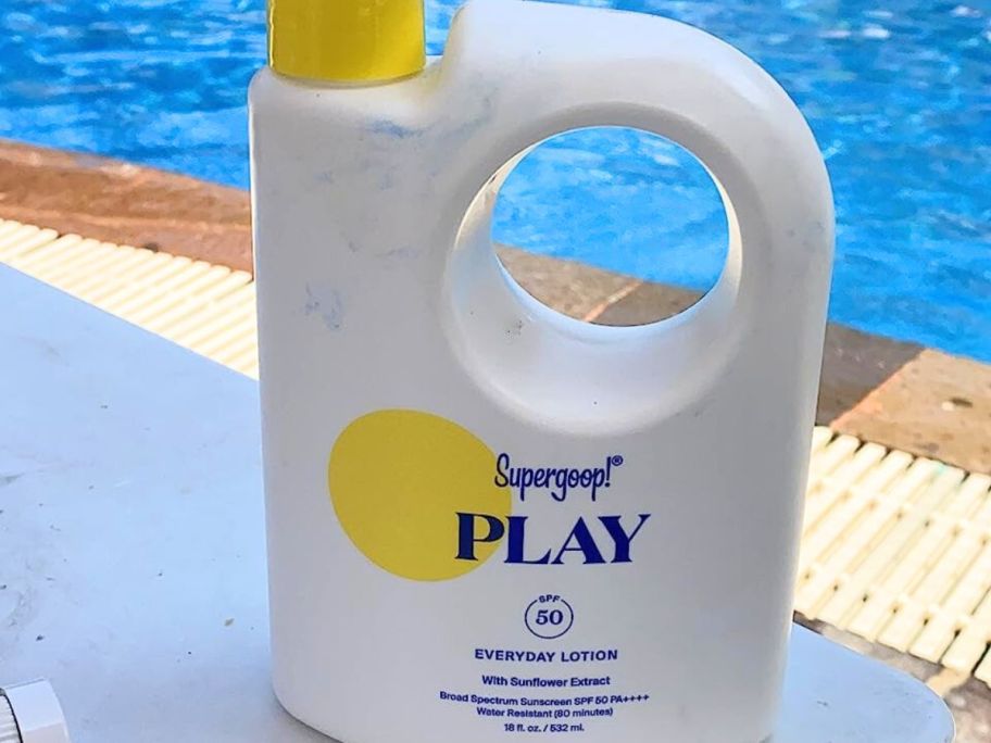 A large 18oz pump bottle of Supergoop play sunscreen on a table by a pool