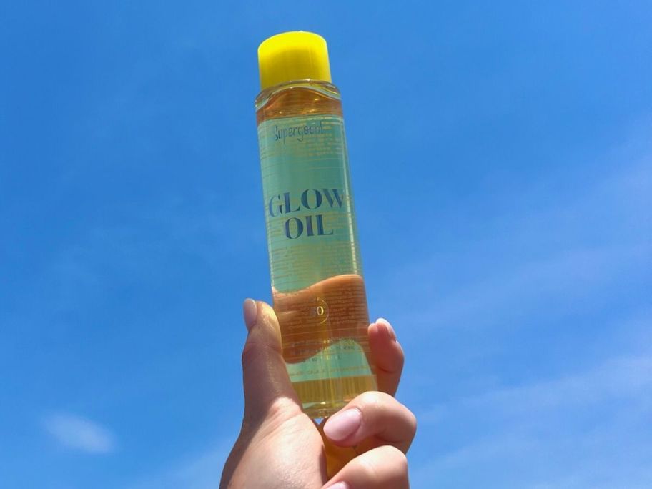 Hand holding a Bottle of Supergoop Glow oil with a blue sky in the background
