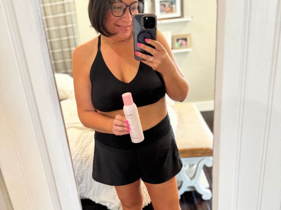 Tan woman taking a picture of herself in a mirror while holding a bottle of Tan Luxe Paris Hilton Spray