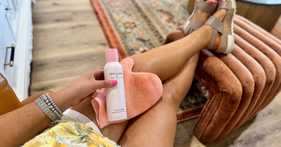 Woman sitting with her tan legs crossed holding a Tan-Luxe Paris Hilton Tanning Spray and Mitt next to them
