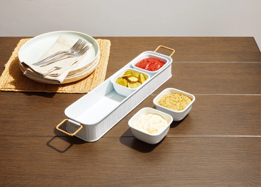 condiment tray with individual cups, both in and out of the tray, on a table near some plates and utensils