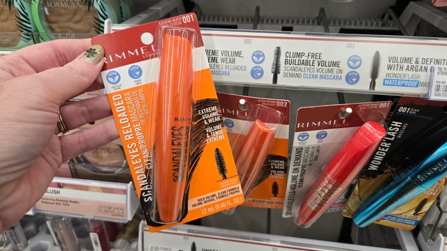 hand holding a rimmel mascara in front of a cosmetics display in a store