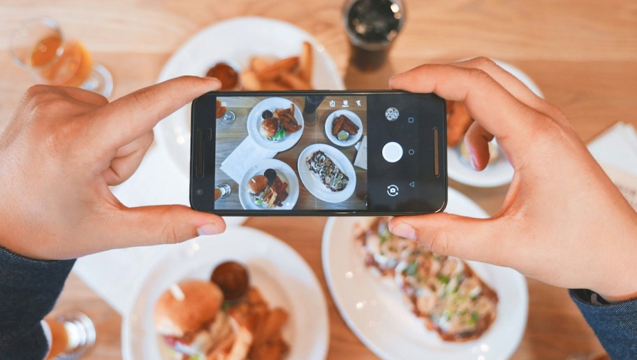 hands holding phone above plates of food taking photo