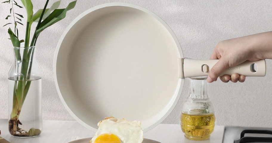 A hand holding a cream-colored Carote nonstick frying pan with a detachable handle, tilting it to slide a fried egg onto a plate.