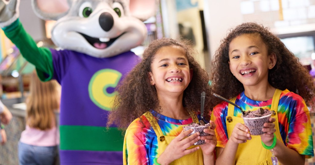Two girls in yellow overalls, holding cups of cookie crunch, dipping dots, and standing in front of Chuck E. Cheese mascot