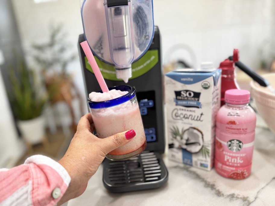 hand holding up a pink frozen drink in front of a slushie machine