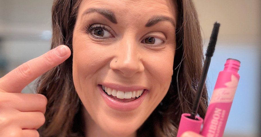 woman pointing to her eye while holding a pink tube of mascara
