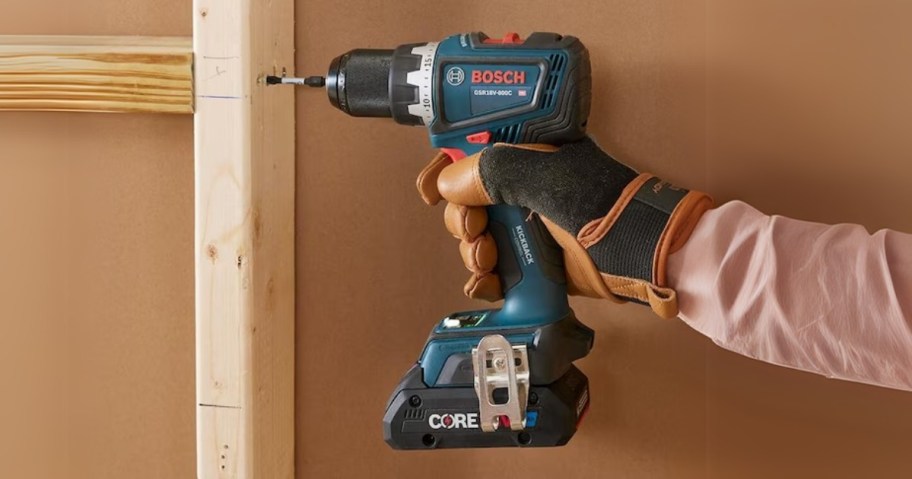 person's hand wearing a glove using a power drill on a wall stud