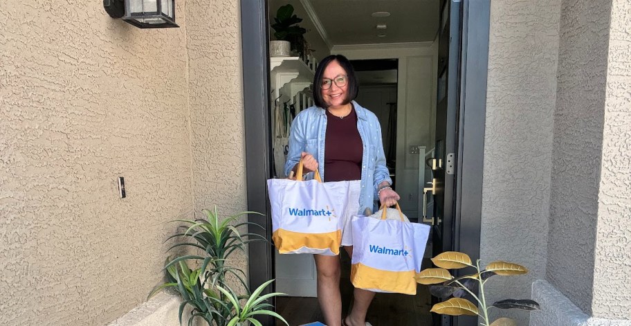 smiling woman standing her in open doorway holding two walmart plus delivery bags