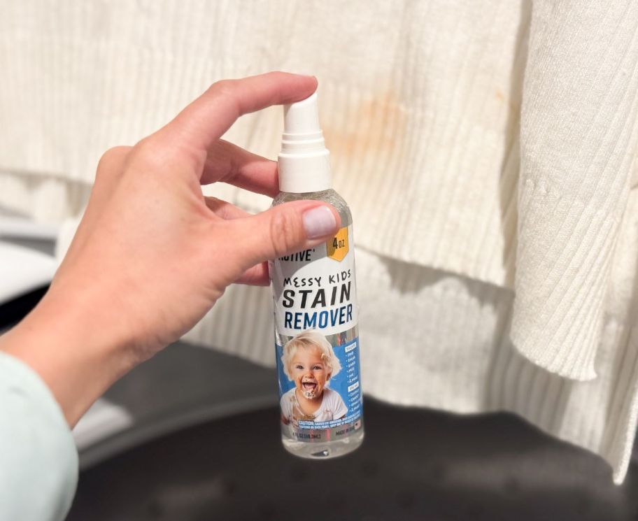 a womans hand holding a bottle of stain remover spray and spraying a stain on a white shirt