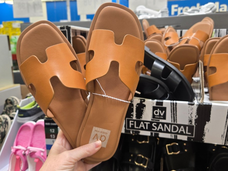 hand holding a pair of women's tan sandals, more tan and black sandals in a box behind them