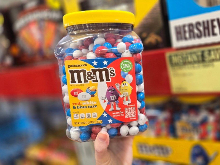 hand holding a large container of red, white, and blue Peanut M&M's in a store aisle