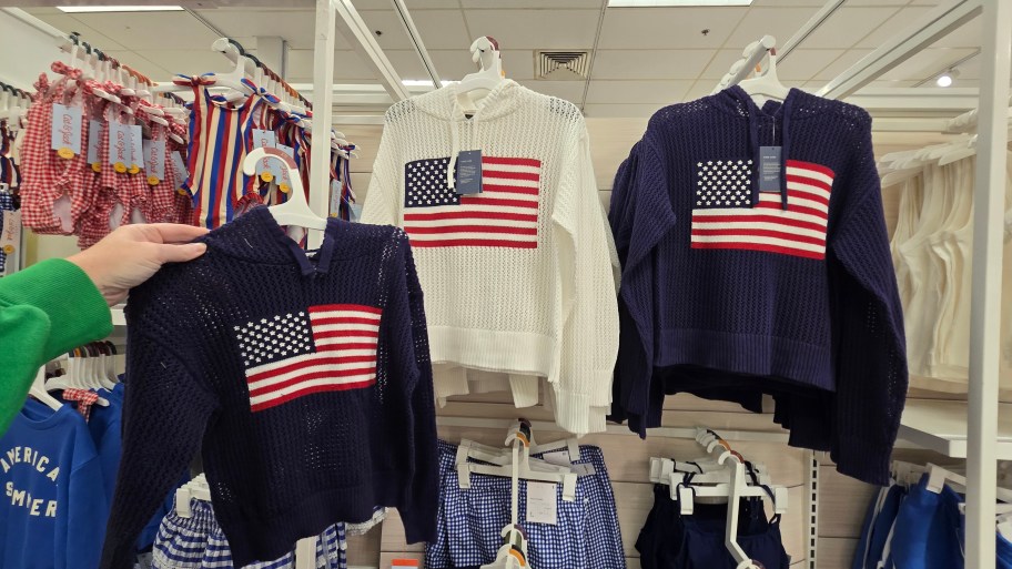 a womans hand holding a navy blue flag sweater up to a white flag sweater hanging on a display wall.. 