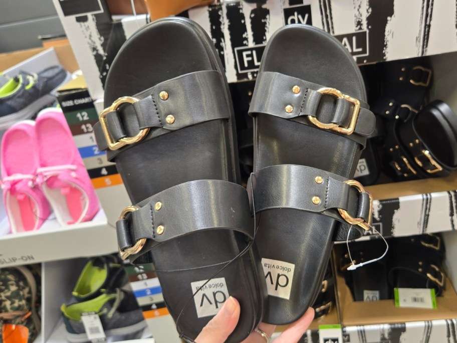 hand holding a pair of women's double buckle black sandals, more in display boxes behind it