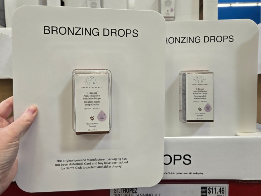 hand holding a package with Drunk Elephant Bronzing drops, more on a shelf behind it