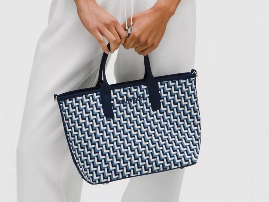 lower half of a woman wearing white pants and holding a knit style small handbag in her hand by her leg