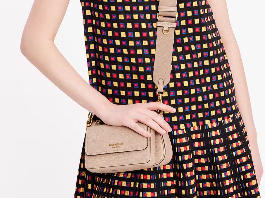 Woman wearing a colorful geometric print dress and holding a beige Kate Spade Double Up Crossbody bag with a wide adjustable strap.